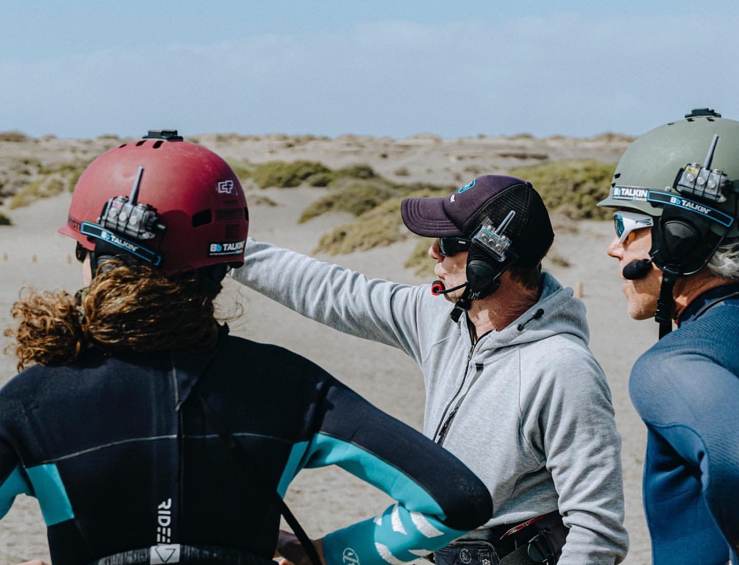 Surf coach standing on the beach briefing three students wearing helmet-mounted BbTalkin intercom headsets before a group lesson