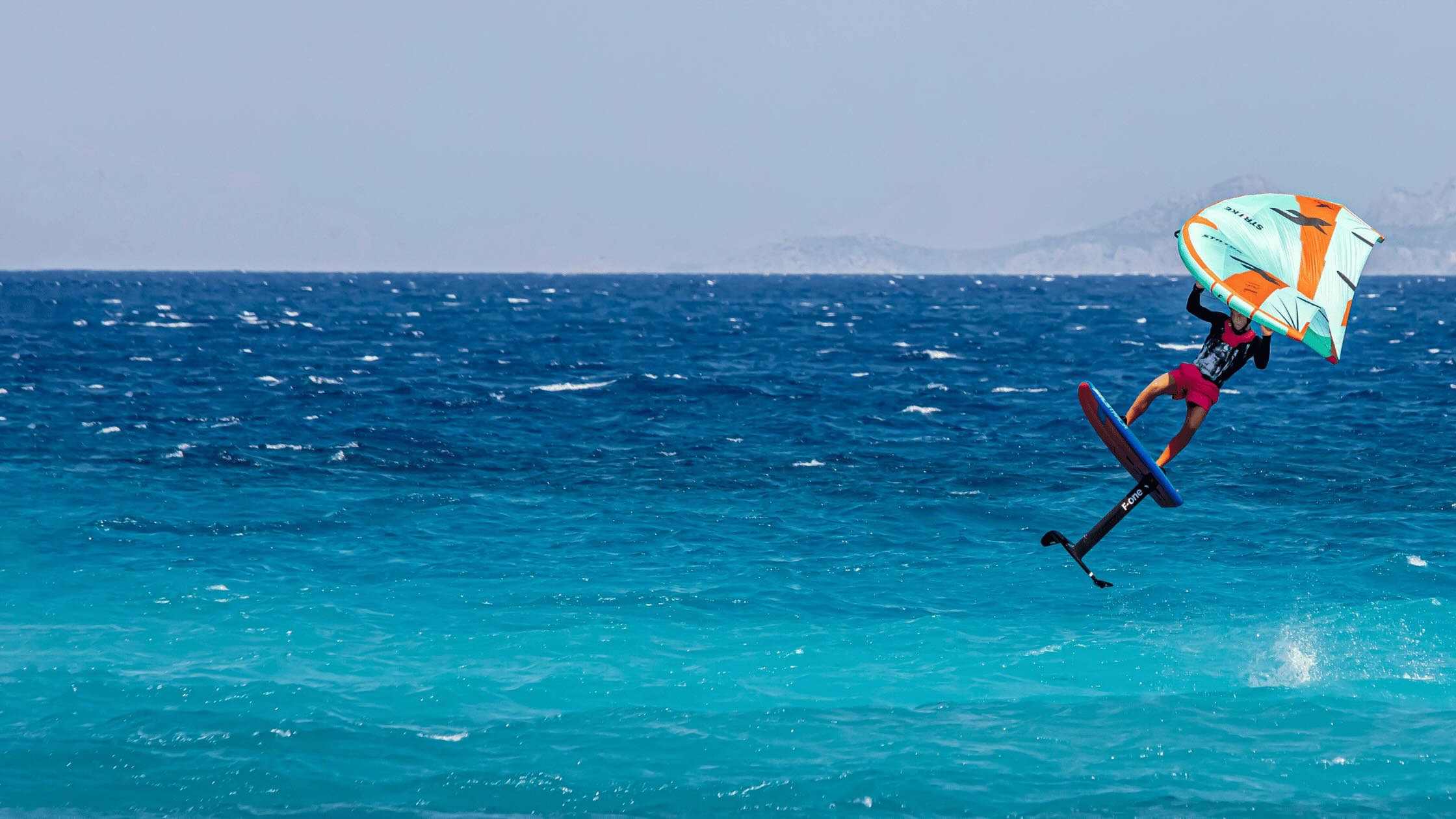 A man on the water e-foiling, up in the air, woth a green and orange kite, over a blue sea 