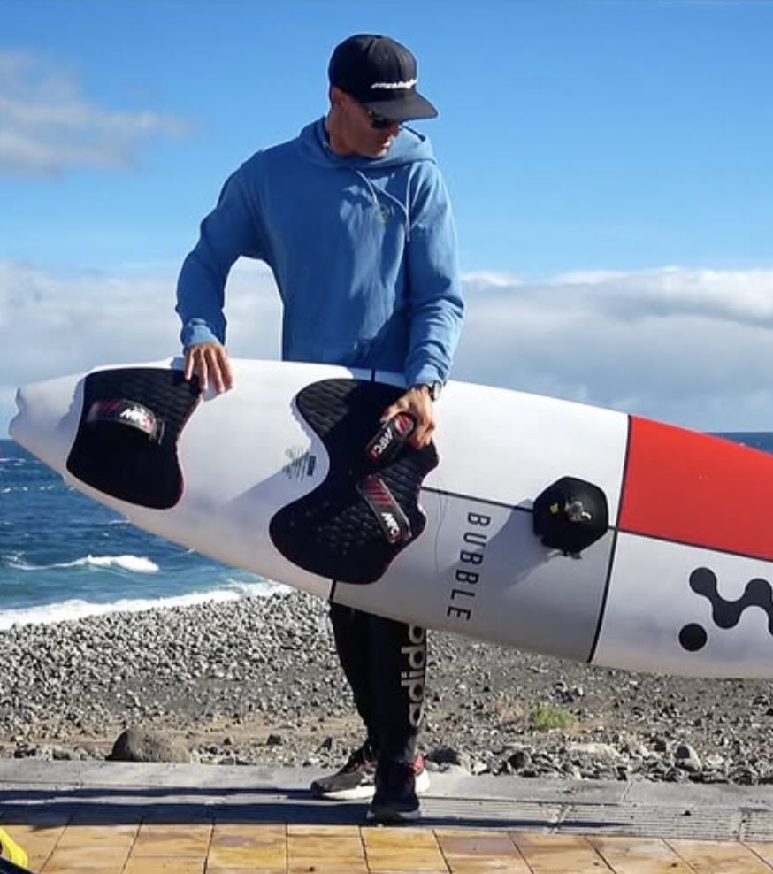 Josep Pons holding a white surfboard with black and red design on a beach