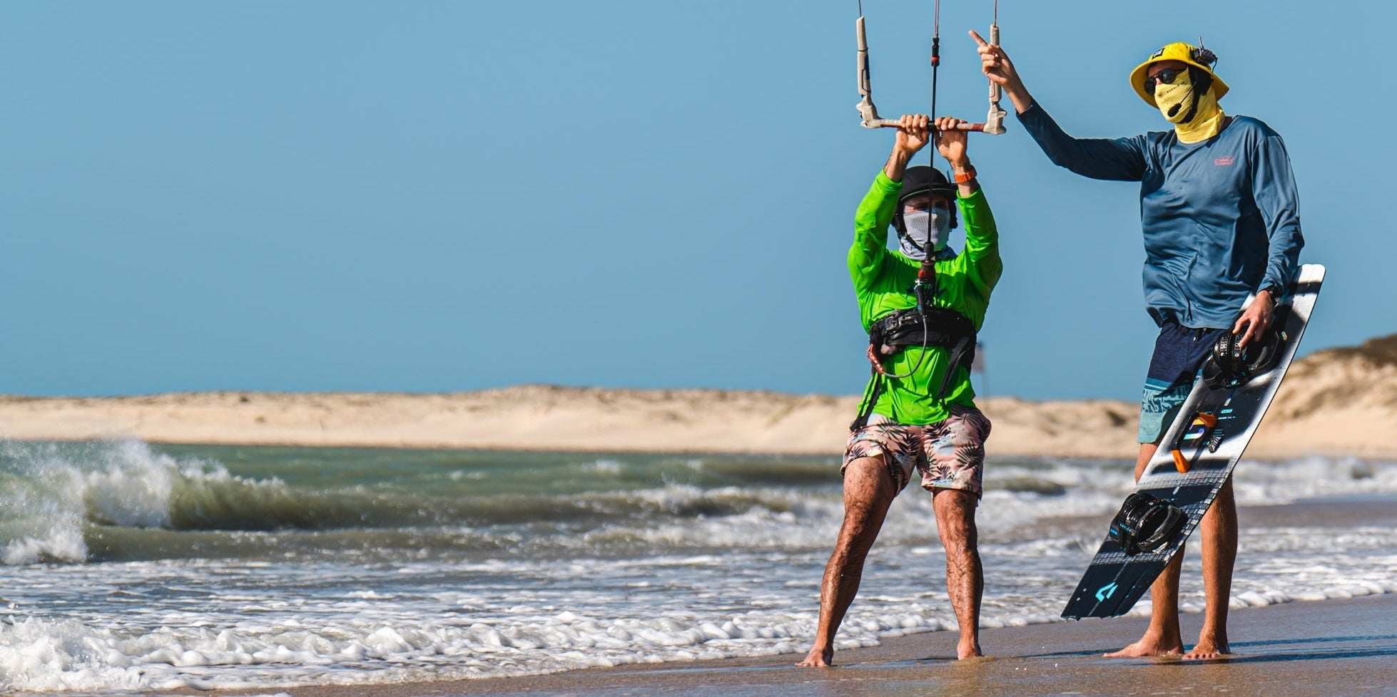 Kite instructor and student standing on a beach with one holding kiteboarding equipment and the other standing on the sand wearing a BbTalkin headset