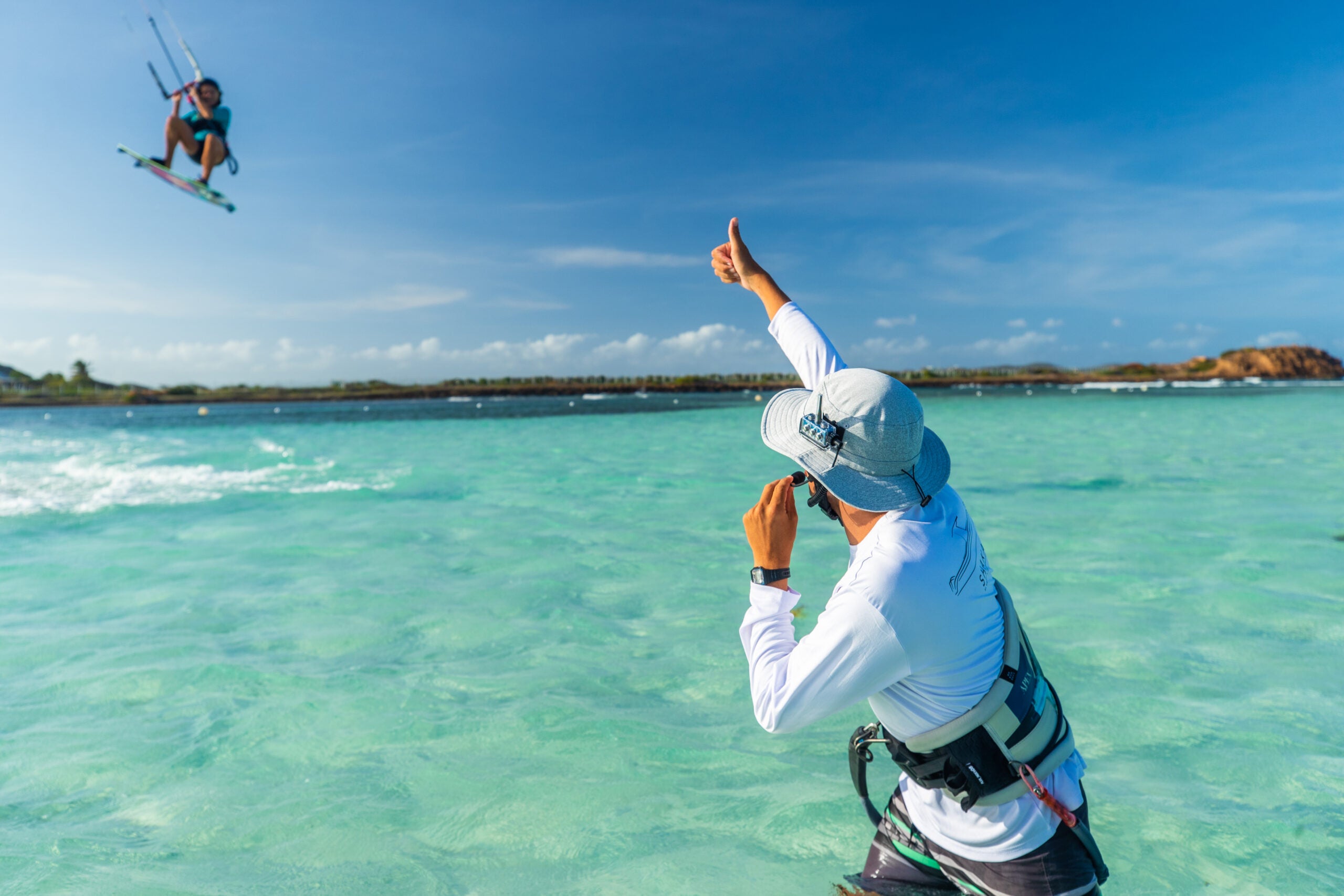 Instructor using a waterproof BbTalkin Bluetooth intercom to coach a kiteboarder jumping over clear blue water, showing hands-free communication for watersports.