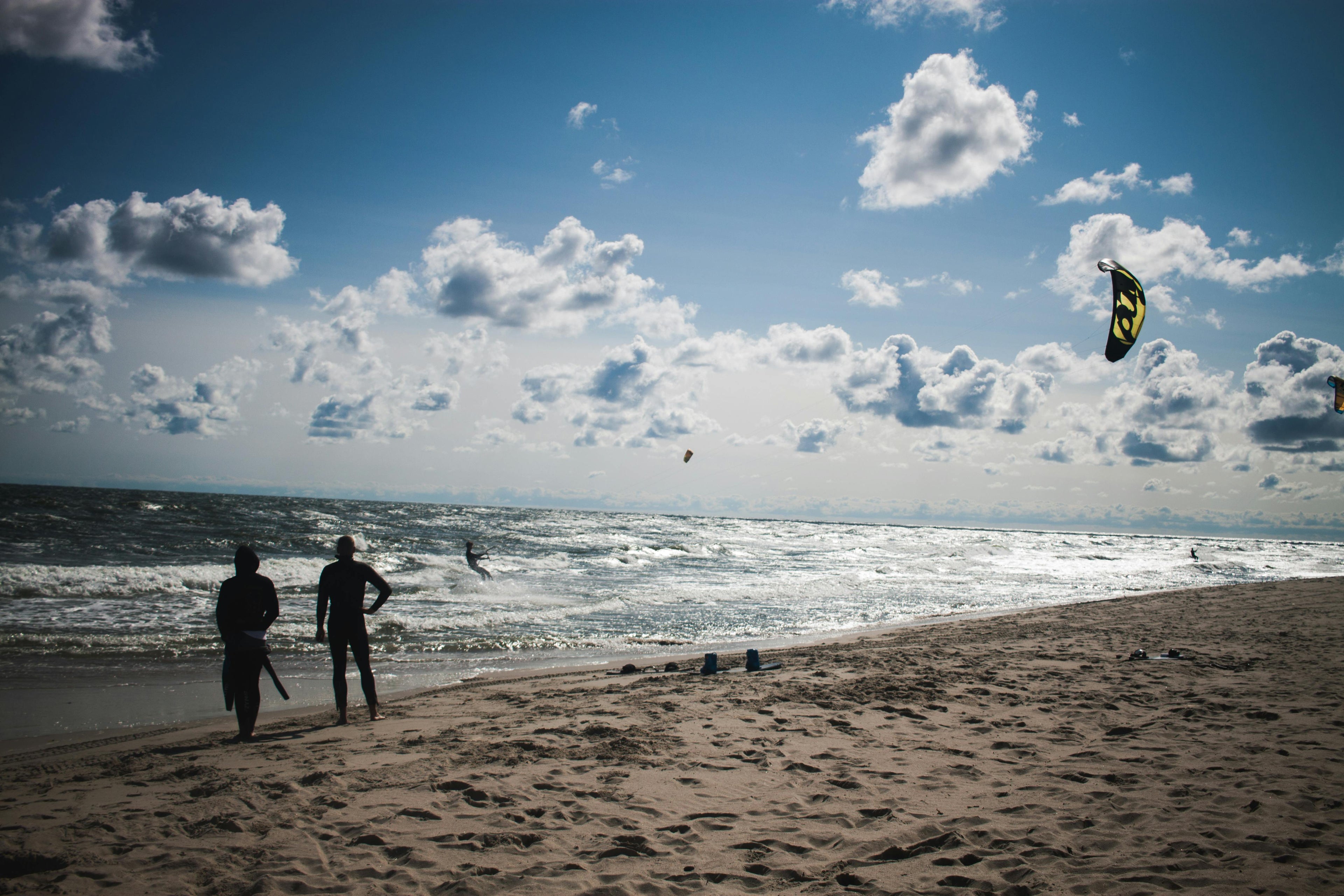 Two people on a beach watchinga. kite surfer, under a blue sky with clouds.